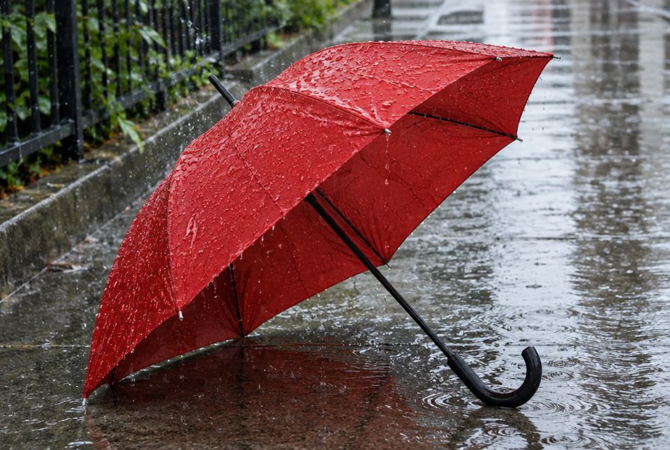 An open red umbrella lying on a wet sidewalk in the rain, covered in raindrops.