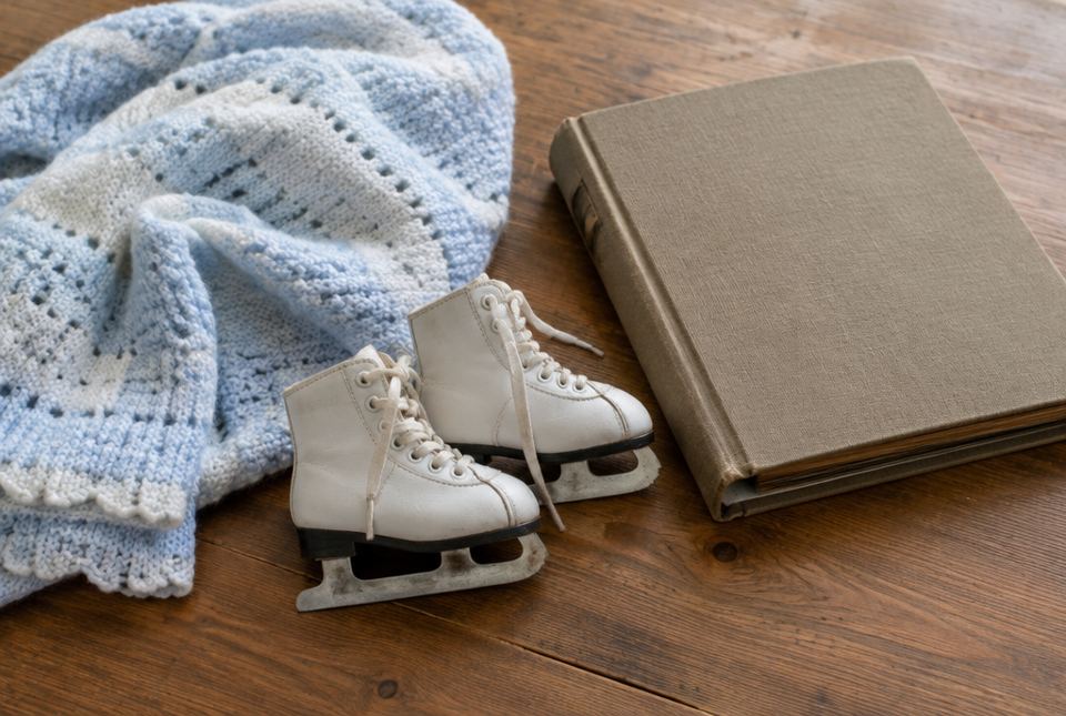 A knitted baby blanket, a pair of small white figure skates and a closed photo album on a wooden table.