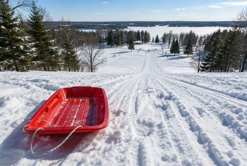 A red plastic sled at the top of a long snowy hill with sled tracks running down to the bottom.