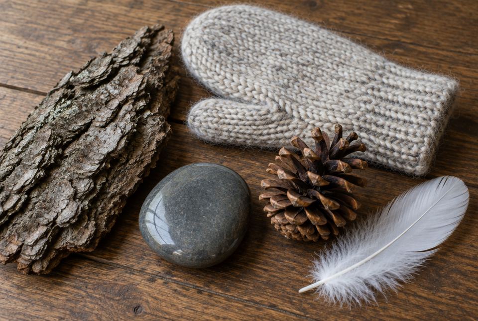 A piece of tree bark, a knitted wool mitten, a smooth grey stone, a pine cone and a white feather on a wooden table.