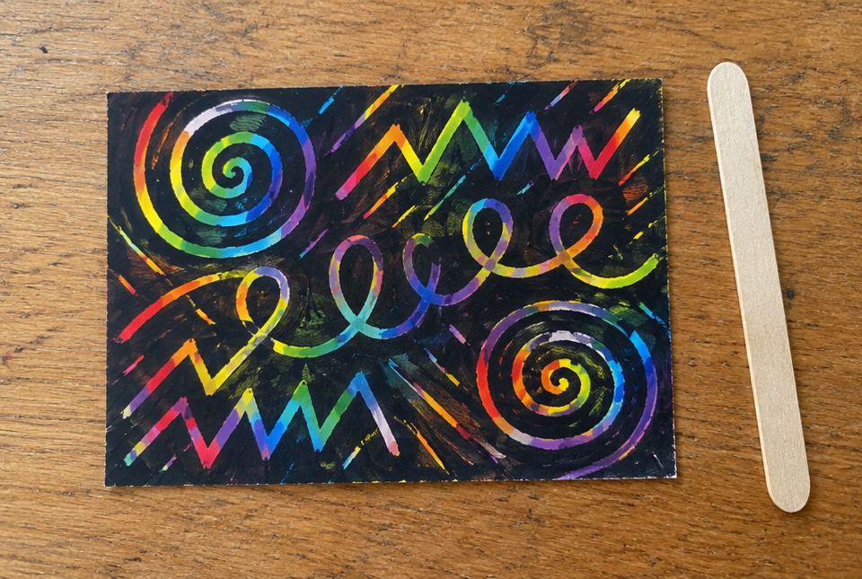 A crayon etching with rainbow spirals, zigzags and loopy lines scratched through black, next to a wooden craft stick.