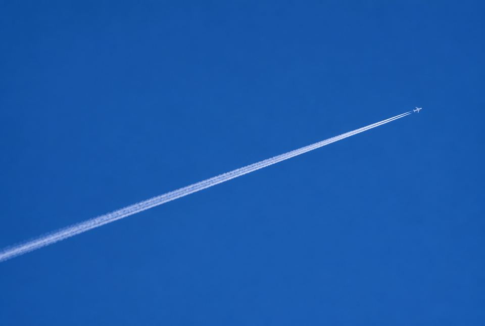 A long white contrail stretching across a deep blue sky behind a tiny, high-flying jet.