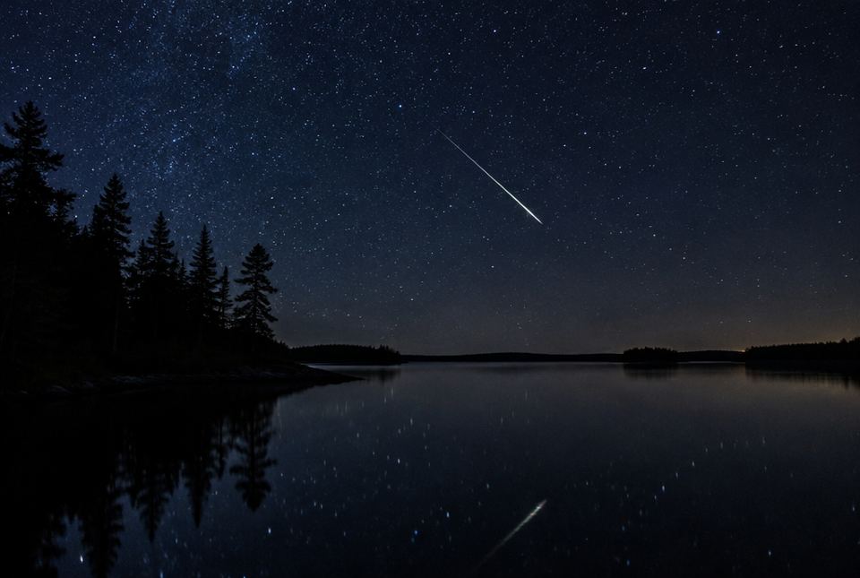 A bright streak of light crossing a starry night sky above a lake and pine trees.
