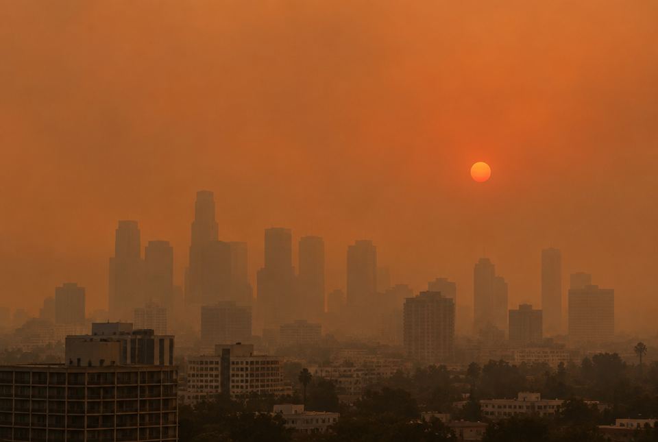 A city skyline under a hazy orange sky, with the Sun a dim red disc through smoke.