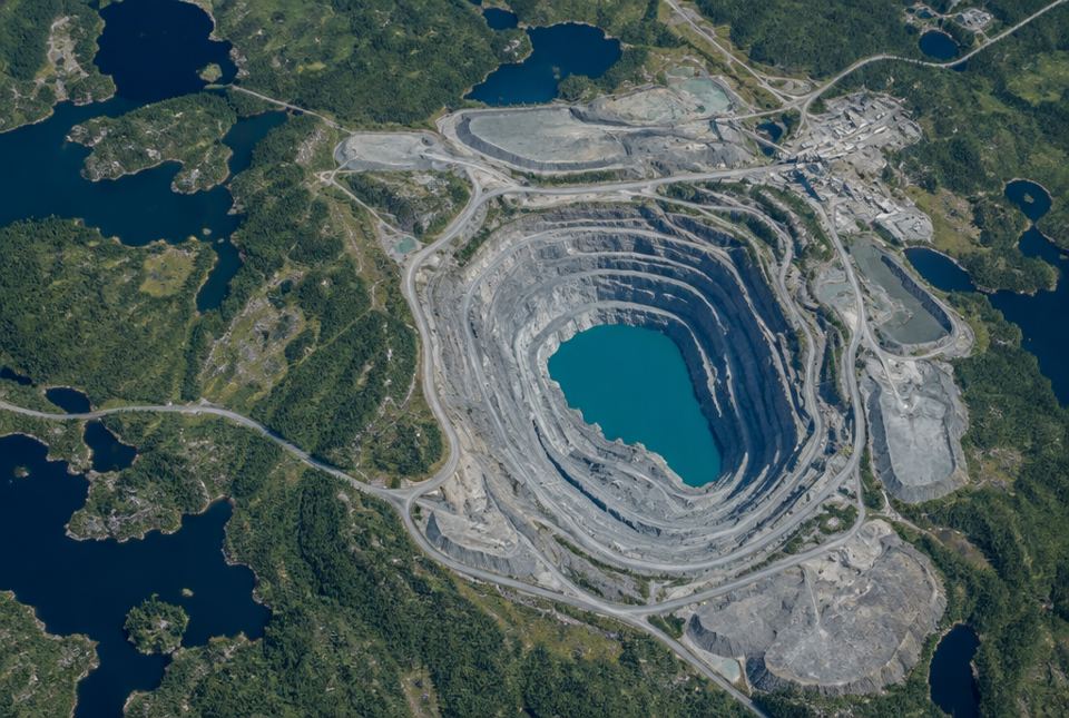 An aerial view of a huge open-pit mine with grey terraced walls and a turquoise lake at the bottom, surrounded by forest and lakes.