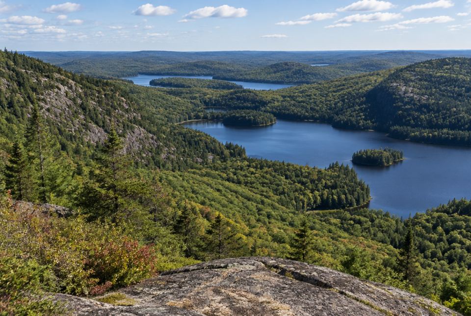 Forested hills and valleys with lakes stretch into the distance below a rocky lookout.