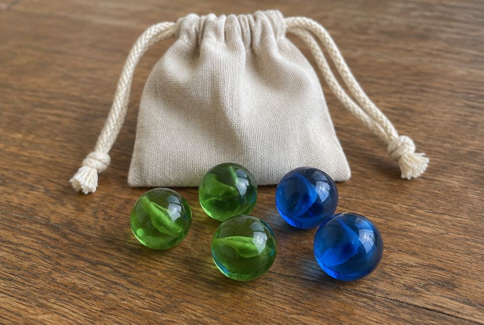 Three green marbles and two blue marbles in front of a small cloth drawstring bag on a wooden table.