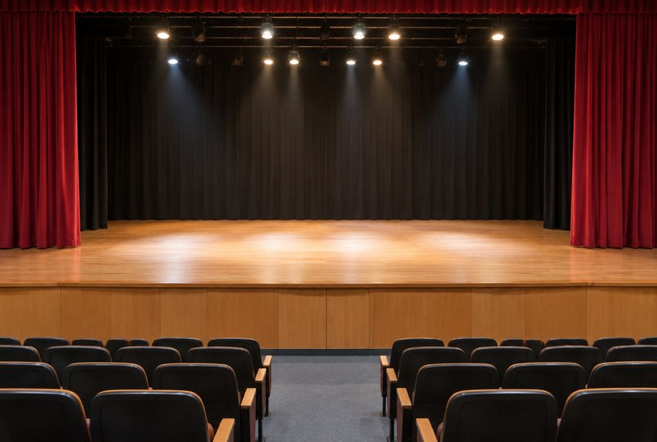 An empty school stage with a wooden floor and red curtains, seen from the audience seats.