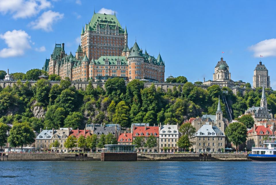 Old Québec City seen from the St. Lawrence River, with old stone houses below a cliff and a large castle-like hotel with green roofs on top.