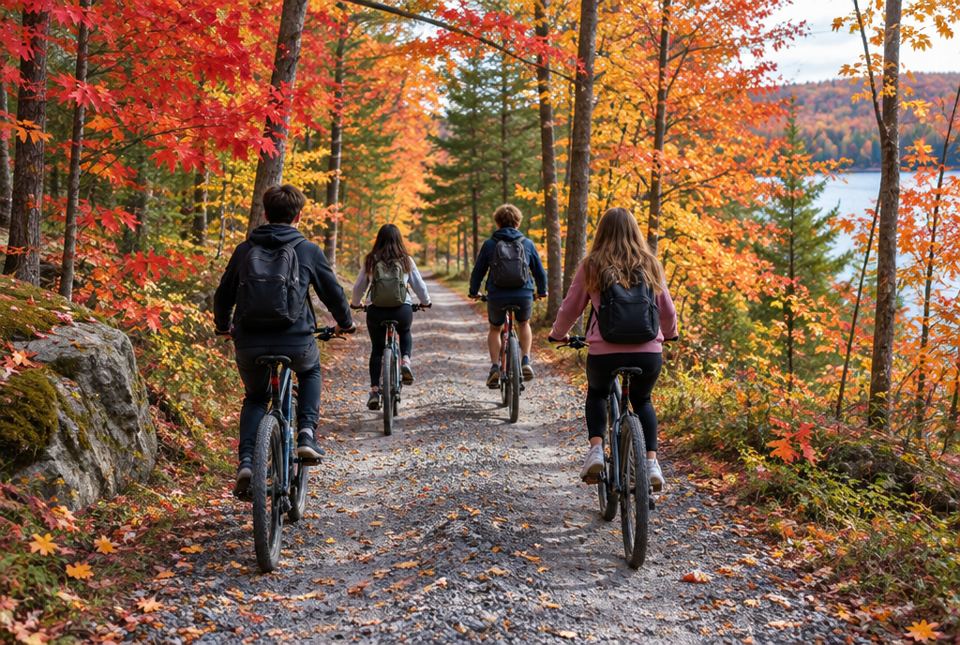 A group of four teenagers, seen from behind, riding bicycles along a gravel trail through a bright red and orange autumn forest beside a lake.