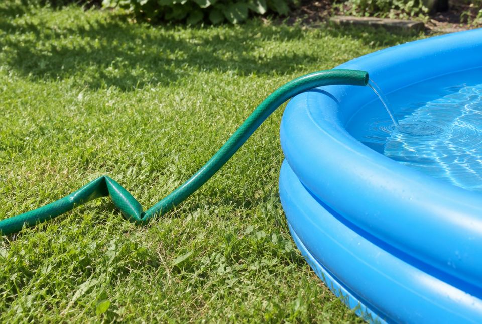 A green garden hose with a sharp kink in it, letting only a trickle of water into a blue wading pool.