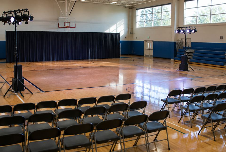 A school gymnasium set up for a performance, with rows of folding chairs facing an open floor and stage lights on stands.