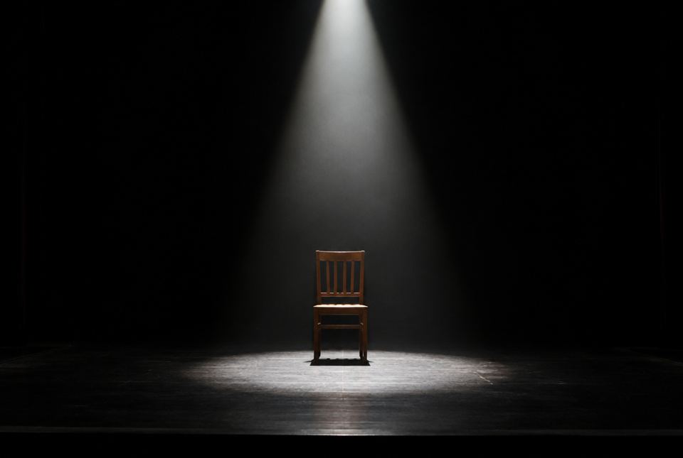A single wooden chair sits in a bright white spotlight in the centre of a dark stage.