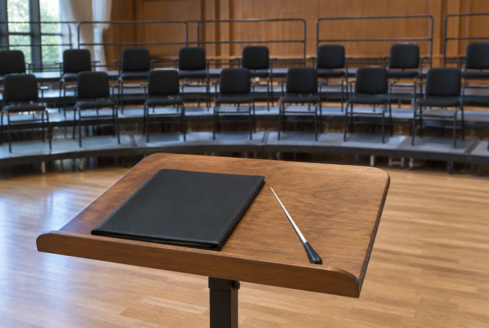 A conductor's white baton and a closed black folder rest on a wooden podium stand facing rows of empty choir chairs on risers.