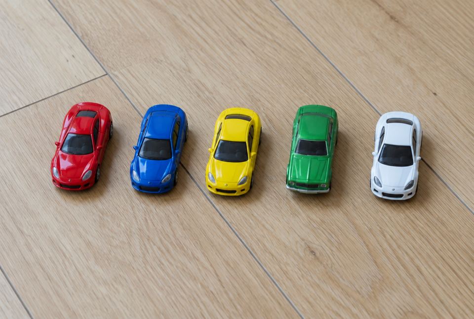Five toy cars in red, blue, yellow, green and white are lined up in a row on a wooden floor.