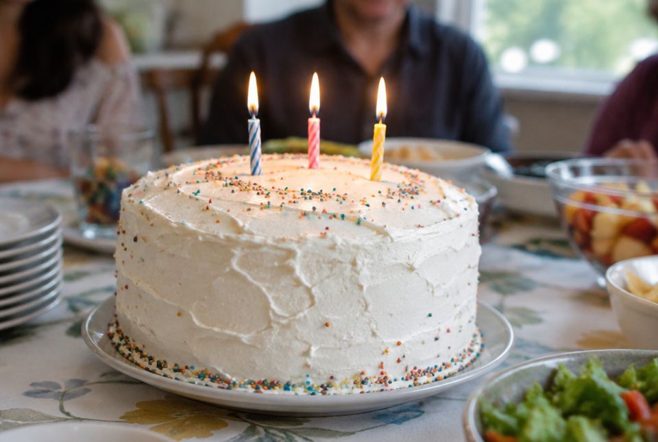 A white birthday cake with sprinkles has three lit candles on top.