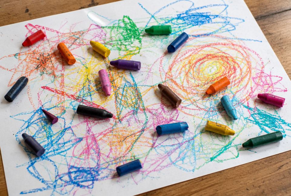 Short and broken crayons in many colours lie on a sheet of paper covered in colourful scribbles.